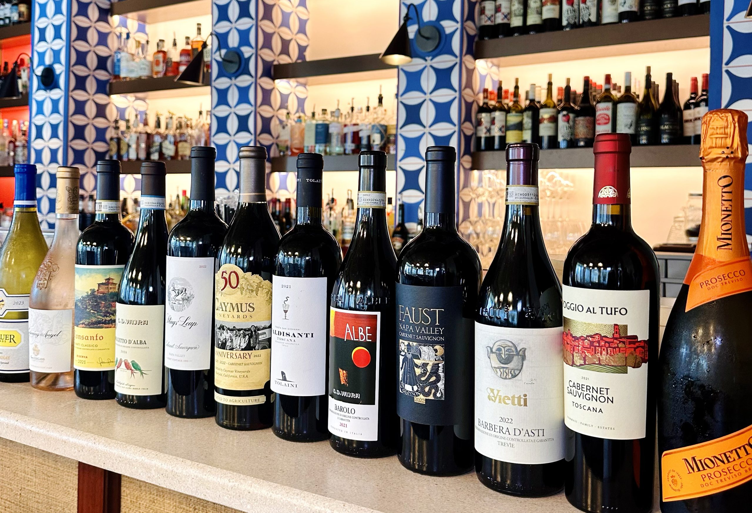 Bottles of Prosecco and Mionetto lined up on a bar against a blue patterned wall, part of Field’s Italian wine list spanning Napa Valley.