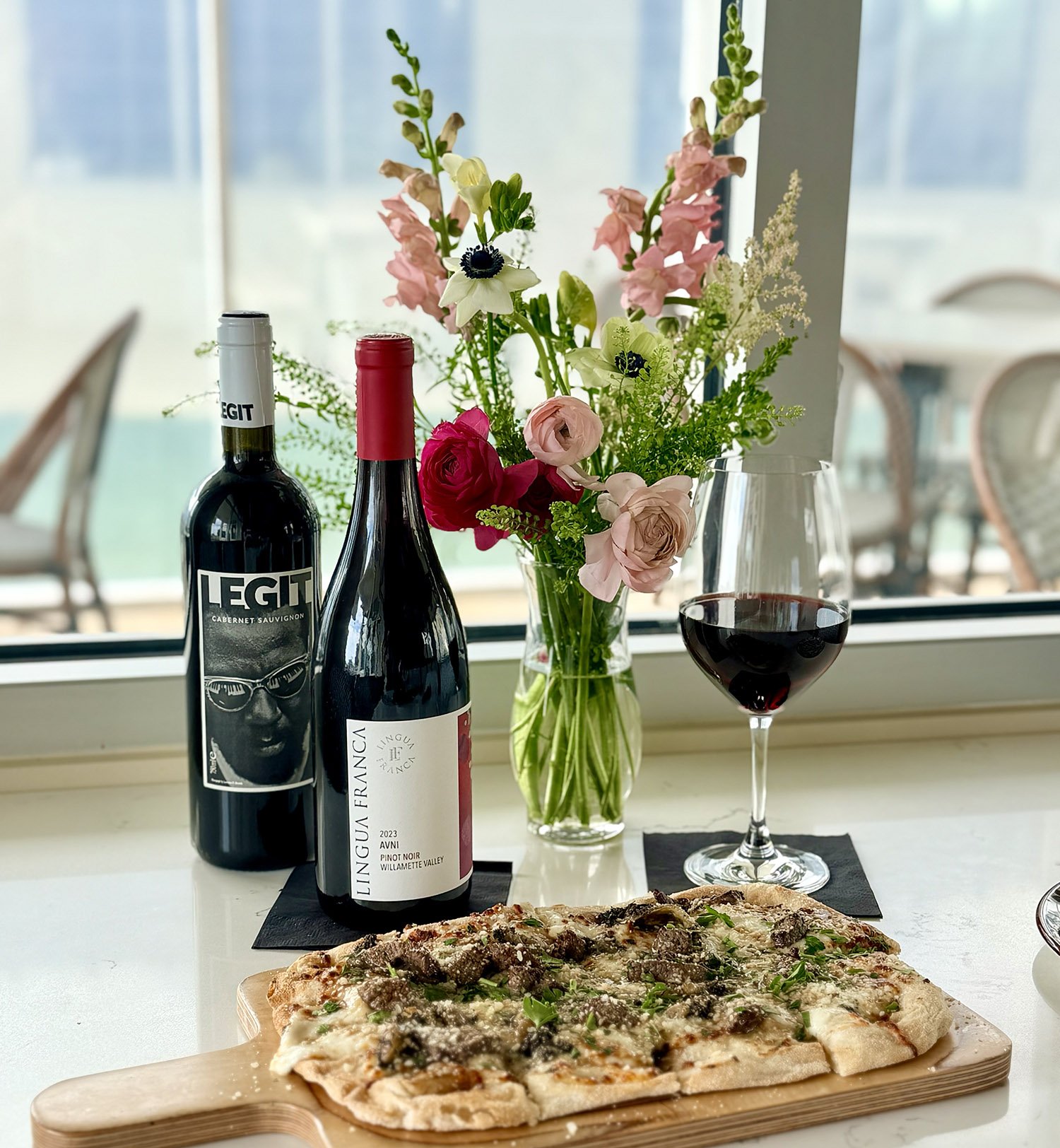 Elegant wine bottles, glasses, and fresh flatbread pizza on the table at Field’s Italian in Pass Christian, with bright pink and green flower bouquet.