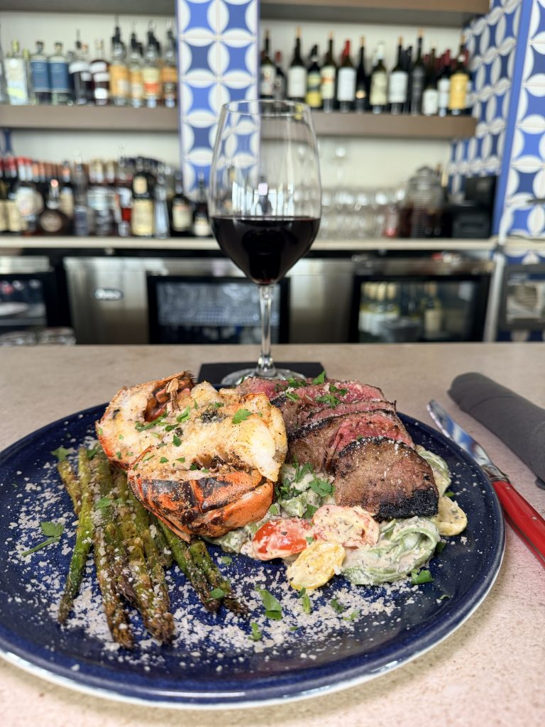 IMG_2764-2 copy Red wine in a clear glass above a plate of grilled steak and seafood with garlic toast, framed by blue-and-white Italian decor at Field’s Italian in Pass Christian.
