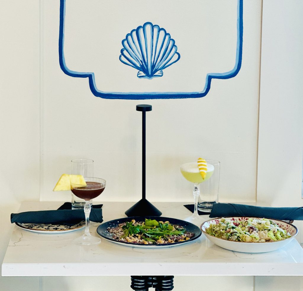 IMG_3329 copy Blue seashell emblem inside a white frame above dining glassware, reflecting Field’s Italian Pass Christian and the coastal Mississippi Gulf Coast spirit.